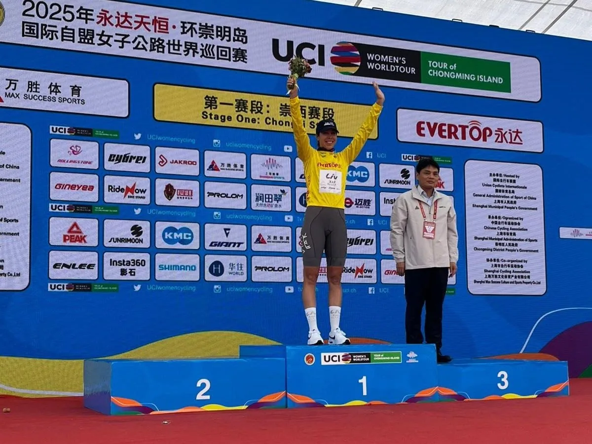 Georgia Baker on the podium at the Tour of Chongming Island, wearing a yellow leader's jersey and standing with her arms aloft in celebration