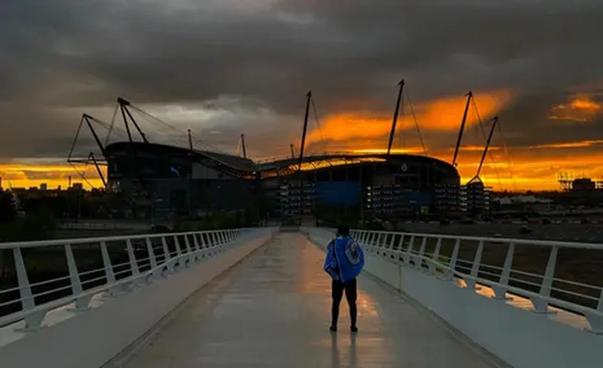 A Manchester City fan watches a golden sunset from a bridge leading to the club's home ground, the Etihad Stadium, behind which the sun is sinking.