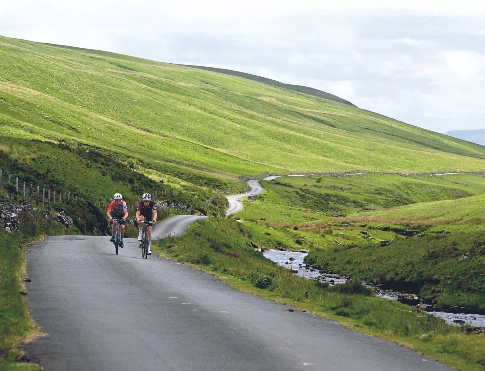 Two riders climb towards the camera with a ribbon like road behind them and fields either side